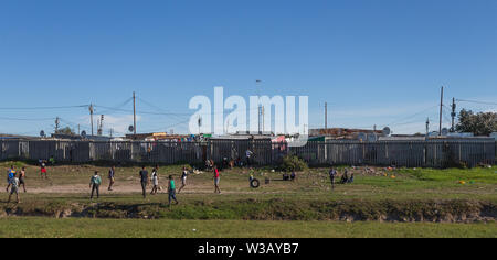 group of poor black African children playing a game of soccer or football on the side of the highway with tin shack houses in the background Cape Town Stock Photo