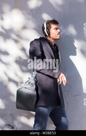 Full length image of business man in eyeglasses and coat smoking cigarette outdoors and listening music with his headphones Stock Photo