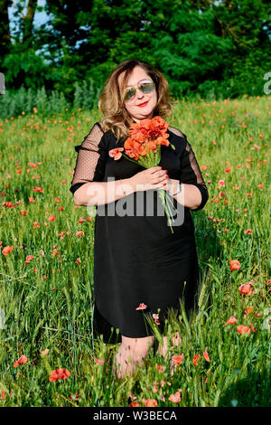 Long-haired plus sized woman in a black dress on a field of green wheat and wild poppies. Overweight fat woman. The concept of freedom and freshness. Stock Photo