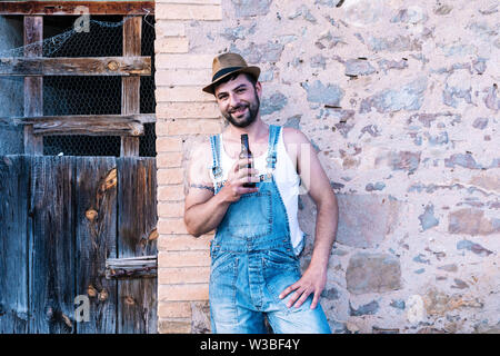 Young bearded man in rural clothing leaning on a wall while smiling and holding a beer Stock Photo