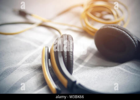 Yellow-black headphones with a microphone, the wire which is tangled in a ball, lie on a gray blanket in a white strip and illuminated by sunlight. Stock Photo