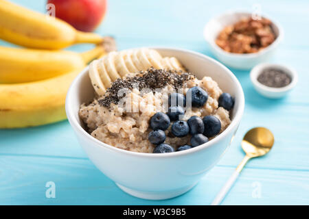 Oatmeal porridge with banana, blueberries and chia seeds in white bowl on blue background. Healthy breakfast food