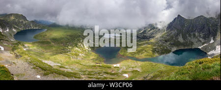 Wide panorama of Rila mountain lakes from a vantage point with dramatic sky, thick mist and smooth, reflective, sapphire blue glacial lakes Stock Photo