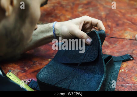 Craftsman intently works with leather. Tanner sews leather goods. Making handmade stuff. Male hands with needle and thread. Man working with needle. Stock Photo