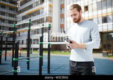 Serious concentrated young man with beard standing on workout area and using smartphone while choosing music for training Stock Photo