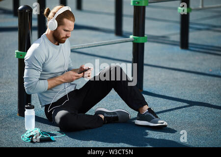 Serious athletic guy with red beard wearing wired headphones sitting on ground and using smartphone while resting Stock Photo