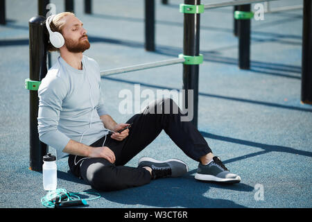 Calm relaxed young bearded man in headphones sitting on sports ground and keeping eyes closed while meditating before training Stock Photo