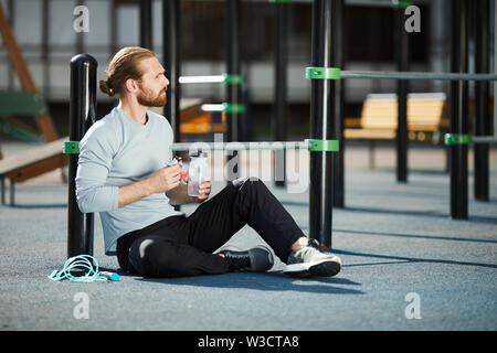 Calm pensive young bearded man in sportswear sitting on public sports ground and eating protein snack and drinking water Stock Photo