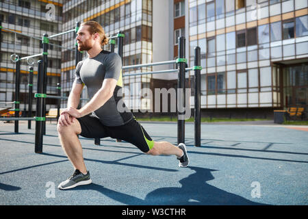 Young red-bearded man in tight sportswear leaning hands on knee and stretching legs while doing static exercise outdoors Stock Photo