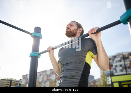 Concentrated determined young bearded guy in tshirt hanging on outdoor bar and lifting own body up Stock Photo
