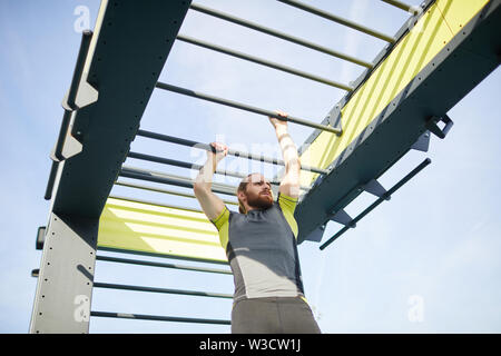 Serious determined bearded man in tight sportswear crossing monkey bars on outdoor training ground Stock Photo