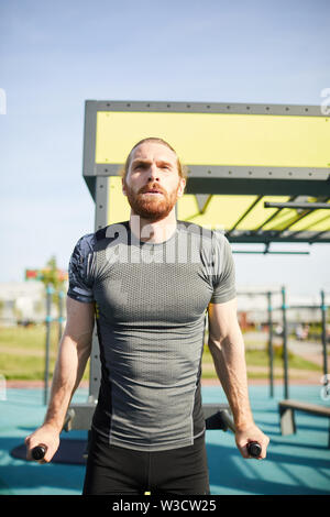 Concentrated tenacious man in sportswear exhaling while doing pull-ups on bars outdoors Stock Photo