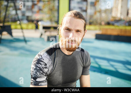 Portrait of satisfied handsome young red-bearded man in sweaty sports tshirt sitting on workout area Stock Photo