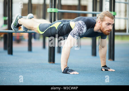 Concentrated determined young red-bearded man in sportswear using sports ground equipment for training outdoors Stock Photo