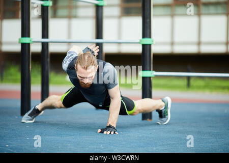 Concentrated strong young bearded man in sportswear doing one armed push up on outdoor workout ground Stock Photo