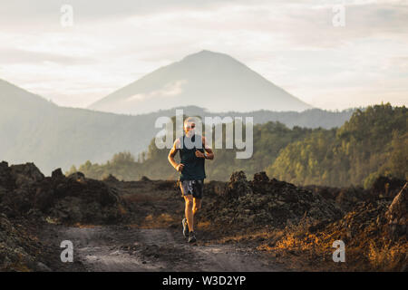 Young athlete man trail running in mountains in the morning. Amazing volcanic landscape of Bali mount Agung on background. Healthy lifestyle concept. Stock Photo