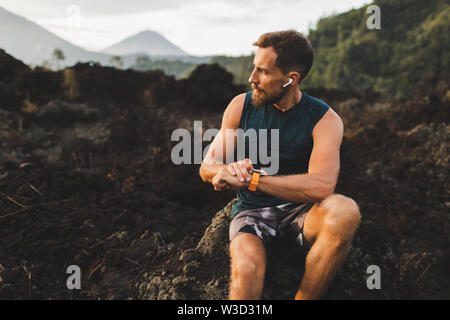 Young hipster runner with beard using smart watch and listening music in wireless headphones before running outdoors. Stock Photo