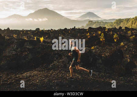 Young male athlete trail running in mountains at sunrise. Amazing black lava volcanic landscape of Bali on background. Adventure sport concept. Stock Photo