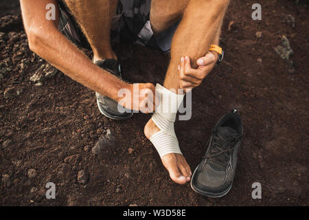 Man bandaging injured ankle. Injury leg while running outdoors. First aid for sprained ligament or tendon. Close-up on dark background. Stock Photo