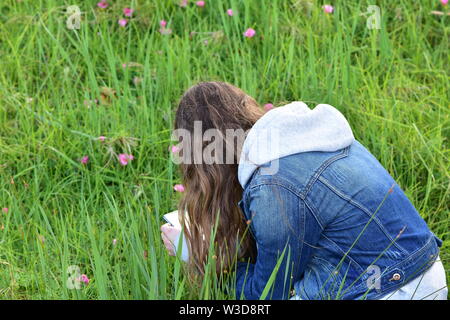 Girl with long hair wearing denim jacket crouching in tall grass among purple flowers. Stock Photo
