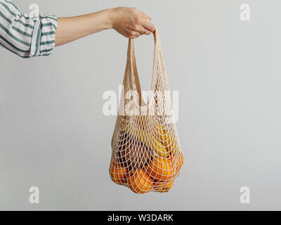 Mesh bag with fruits in female hand. Stylish young woman hand hold mesh shopping bag on light gray wall. Modern reusable shopping concept. Stock Photo