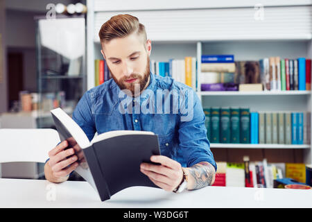 Studying for an exam. Focused handsome young man studying from textbook and sitting at table in college library Stock Photo