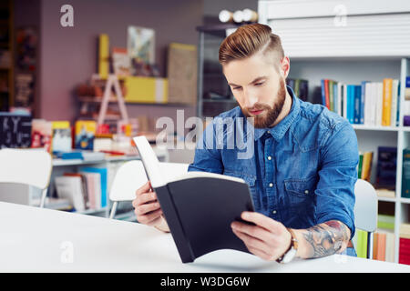 Concentrated handsome young man reading textbook while sitting in library Stock Photo