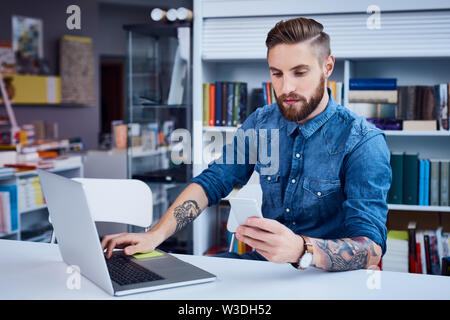 Young man, hipster using his smartphone and laptop in library Stock Photo