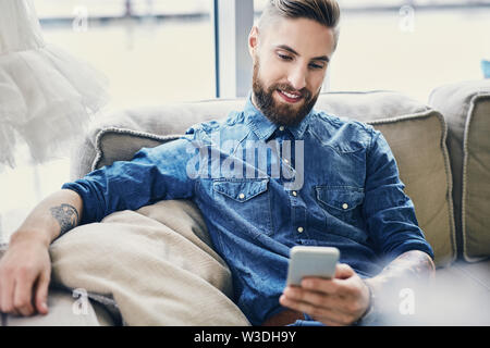 Happy tattooed man using his smartphone sitting sofa Stock Photo