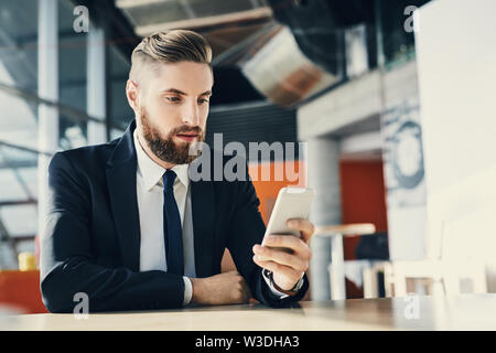 Young man in suit sitting by table browsing his smartphone at modern office Stock Photo