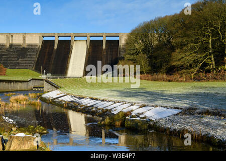 High, steep, concrete dam at Thruscross Reservoir reflected in water flowing in channel on frosty winter day (blue sky) - North Yorkshire, England, UK Stock Photo