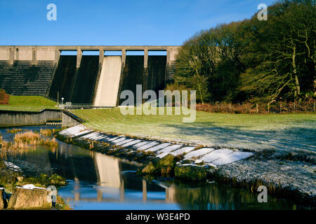 High, steep, concrete dam at Thruscross Reservoir reflected in water flowing in channel on frosty winter day (blue sky) - North Yorkshire, England, UK Stock Photo