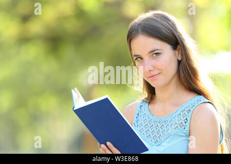 Serious woman looks at camera holding a blank paper book on green background in a park Stock Photo