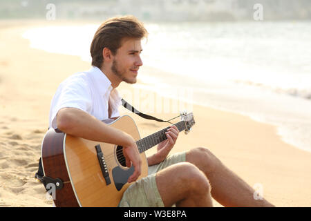 Side view portrait of a relaxed man playing guitar sitting on the sand on the beach Stock Photo