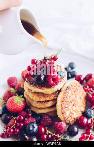 Woman pouring maple syrup on tasty cottage cheese pancakes served with lots of fresh berries on white background. Stack of Pancakes with pouring honey Stock Photo