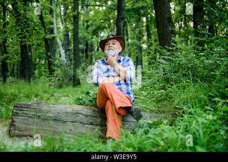 thoughtful man. hiking in deep wood. forest owner. summer or spring picnic. senior man farmer relax in forest. mature man with beard in hat. farmer sit on wood. happy forester. human and nature. Stock Photo