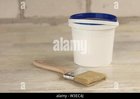 A paint brush is next to a plastic paint bucket with a blue lid on an old white vintage wooden board. Place for text or logo. Stock Photo