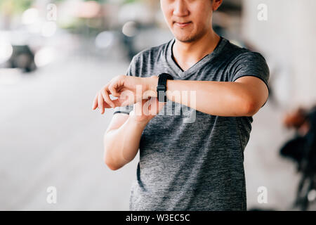 Male in gray T-shirt checking fitness tracker while standing on blurred background of city street during outdoor workout Stock Photo