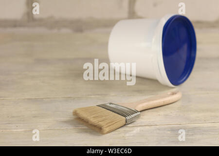 A paint brush is next to a plastic paint bucket with a blue lid on an old white vintage wooden board. Brick wall in the background. Place for text or Stock Photo