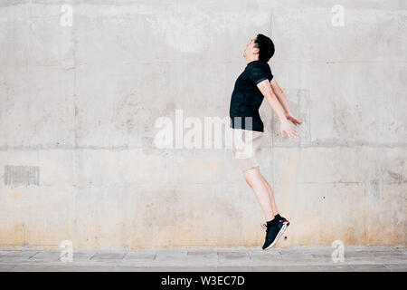 Side view of Asian male in sportswear jumping in the air against shabby wall during outdoor workout on city street Stock Photo