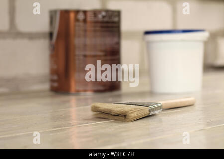 A paint brush is next to a plastic paint bucket with a blue lid and metal can on an old white vintage wooden board. Brick wall in the background. Plac Stock Photo