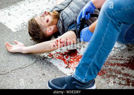 Applying first aid to the injured bleeding man, wearing tourniquet on the arm after the road accident on the pedestrian crossing Stock Photo