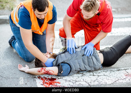 Medic with car driver applying first aid to the injured man lying on the pedestrian crossing after the road accident Stock Photo