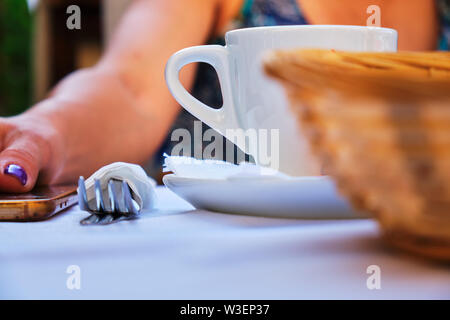 Breakfast, lunch, or dinner concept with a white cup of coffee, a fork, and a bread basket, all set up in front of a woman putting down her phone on t Stock Photo