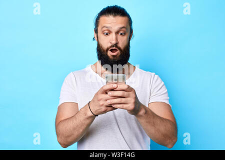 shocked young strong muscular man looking at mobile phone isolated over blue background. man is being shocked to receive sms. close up portrait. studi Stock Photo