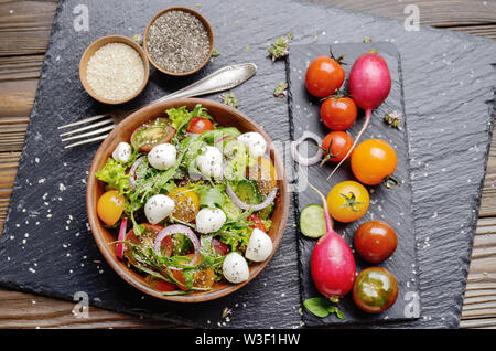 Flat lay of vegetable salad with mozzarella cheese, lettuce, cherry tomatoes, radish, cucumber, onion and basil in clay dish Stock Photo