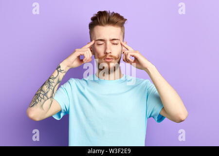 Young relaxed man concentrating on thoughts,suffering from headache, pressing fingers to temples, isolated on blue background. relaxation, man massagi Stock Photo