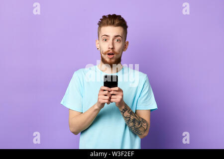 handsome surprised puzzled young man in blue t-shirt, holding smartphone, looking scared and confused reading text message. isolated blue background. Stock Photo