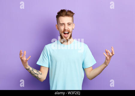 angry young man with moustache in blue shirt standing with raised arms and looking at camera, shouting and screaming. indoor studio shot isolated on b Stock Photo