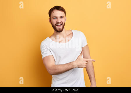 Happy excited bearded man in stylish white T-shirt pointing away at copy space with finger isolated over yellow background, direction, this way. look Stock Photo